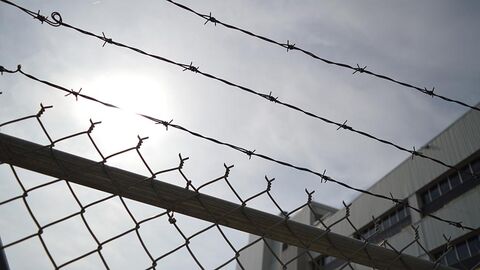 Zaun mit Maschen- und Stacheldraht, im Hintergrund ist ein Teil eines Beton-Gebäudes zu sehen.