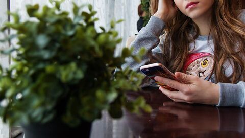 Junges Mädchen Junges Mädchen sitzt an einem Tisch und hält ein Mobiltelefon in einer Hand.